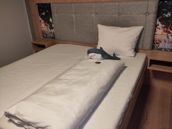 A neatly made bed with a gray upholstered headboard. A single white pillow and a plush shark toy are positioned on the bed, which is covered with a white duvet. A nightstand made of wood is visible beside the bed.