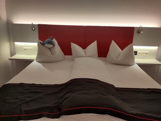 A cozy hotel bed with two large white pillows, a black blanket with a red stripe, and a blue stuffed shark toy resting on one of the pillows. The headboard is red, and there are modern lamps and a nightstand with outlets nearby.