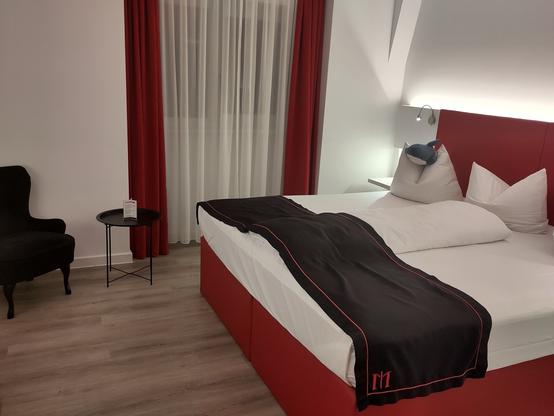 A modern hotel room featuring a large bed with white bedding and decorative pillows. A black throw blanket is placed on the bed. There is a small black side table next to a stylish black armchair. The window has sheer white curtains and red drapes. A plush shark rests on top of one pillow.