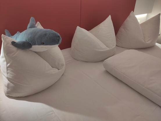 A plush shark toy is perched on top of two large, white, triangular pillows on a bed with a plain white blanket. The background features a red wall.
