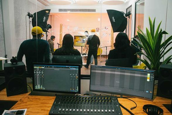 Fotografía. Cabina de grabación de videopodcast, mesa con consola y pantallas en primer plano. Al fondo se encuentra un cuarto insonorizado con seis personas.
