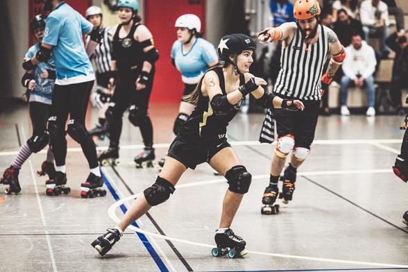 A color picture of a Roller Derby game. A jamming person is coming round the corner with the jammer ref behind them.