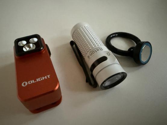 Nahaufnahme: Zwei Taschenlampen von Olight und ein runder Karabinerringer mit Magnet liegen auf einer weißen Tischfläche.