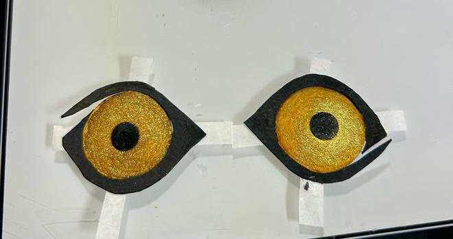 Detail of the construction of the eyes. They are made of punched circles dyed with gold ink with small black circles dyed with acrylic ink for the pupils. Black watercolor paper has been cut into the eye shape. Strips of deli paper have been used in a cross behind the “eyeballs” to affix them to the black paper.