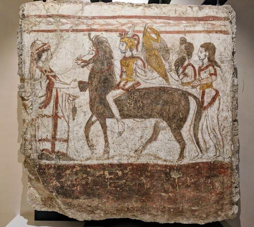 A fresco from Paestum depicts a male figure riding a horse in the center, with a person on the left and two figures on the right, painted on a white background with a rough, plastered border. Apparently, it once decorated the walls of an ancient Lucanian tomb in Paestum, dating to around the 4th century BC, and depicts the return of the warrior.