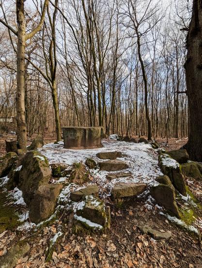 Das Foto zeigt eine Waldszene mit einer Steinstruktur in der Mitte. Die Steinstruktur ist ein rundlicher Altar, umgeben von einem Ring aus großen Felsen. Es gibt Steinstufen, die zum Altar führen, und der Boden ist teilweise mit einer leichten Schneeschicht bedeckt. Die Bäume im Hintergrund sind hoch und blattlos. Die Szene wirkt friedlich und hat einen alten, mystischen Charakter.