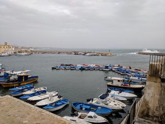 A high-angle shot depicts a harbour filled with numerous small boats, with a town and breakwater in the background under an overcast sky.