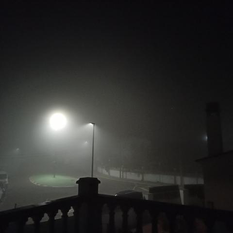 Fotografía nocturna d'una escena envolta en una densa boira. Un fanal il·lumina un àrea circular en primer pla, una part d'una barana en la part inferior de la imatge. Altres faroles són visibles a la distància, però estan enfosquides per la boira.