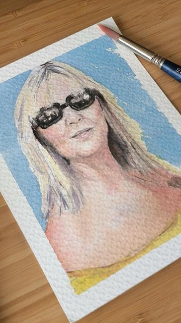 Photo of a watercolor on a desk next to a paintbrush, in the watercolor you can see a blonde woman with sunglasses on a light blue background