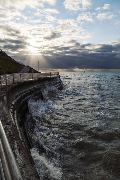 We're on a promenade next to fairly rough sea and sunset is almost upon us. The promenade curves from under our feet to the left out of view then back in to a rounded point directly ahead of us where a narrow paved ramp leads up the hillside we can just see to the left and where the promenade continues around out of sight. Metal railings protect us from the tapered, 2-metre high sea wall of ribbed-looking concrete. Clouds are gathering on the horizon with the onset of evening. A thin band of clear sky at the base on the horizon allows a warm glow from the sun mostly behind the clouds to cast some heat. Where the sun is we can see bright white and long sun beams stretching out in all directions over the grey cloud face to reach the sea. The patchy, clearer sky to the top right is a pale blue. The water appears a green-black ink, textured with flecks of foam on its waves. Water has crashed with enough force against the sea defences ahead of us to send three white jags of waves upwards seeking access to the promenade. It feels powerful and the weak sunshine is welcome against what must be a chilly breeze coming off the sea.