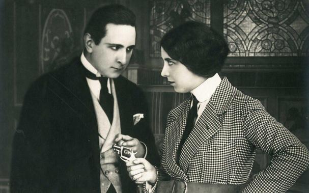 In a black and white film still, a woman points a small pistol at a man, the two staring at each other in early 20th century fashion.