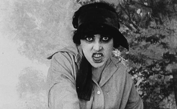 A black and white frame of a menacing young woman, black bags under her eyes, snarling at the viewer.
