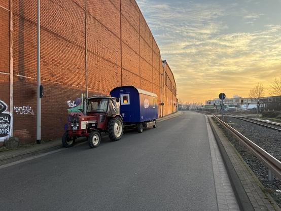 A youth project tractor with a trailer in front of a long read brick wall in the golden glow of the setting sun.