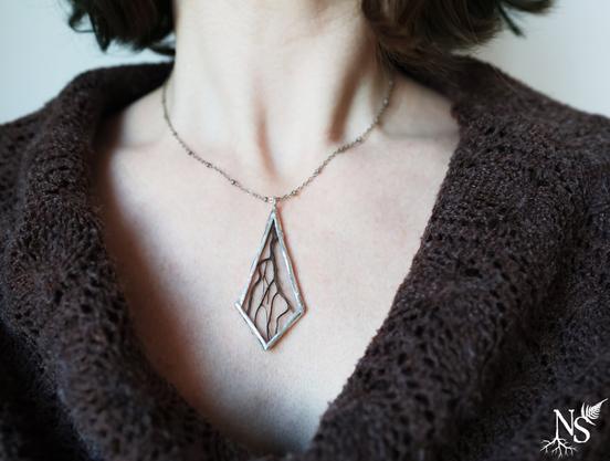 English A woman's neckline with a brown crochet top and a simple silver necklace with a stained glass pendant containing real roots bits.
❉
Français Un décolleté de femme avec un haut en crochet marron et un collier simple en argent avec un pendentif en vitrail contenant de véritables bouts de racines.