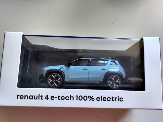 A small model of a light blue SUV, labeled as "Renault 4 e-tech 100% electric," is displayed inside a transparent box. The model showcases details such as the wheels, windows, and body lines of the vehicle. The box bears the name and specifications of the model along the bottom. The background is plain, emphasizing the model.