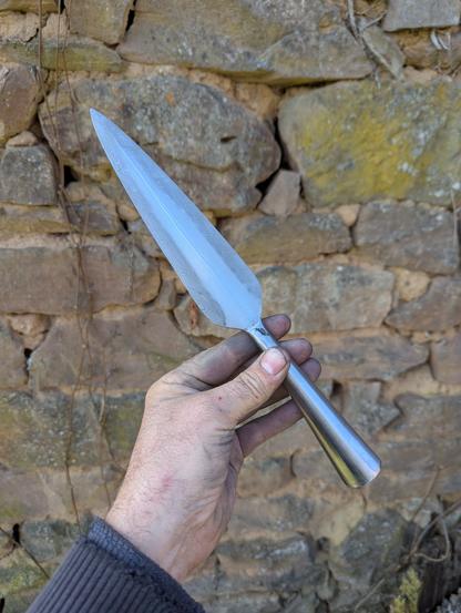 sur un fond de mur en pierres, ma main qui tient la pointe de lance par la douille, elle est en acier, on voit bien le tranchant rapporté en damas.