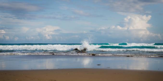 You can see the shore of a beach from a low point of view and how a wave breaks lightly when it reaches the shore. In the background a blue sky with light clouds.
