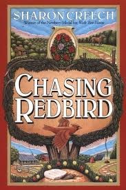 Chasing Redbird by Sharon Creech. The title is centered on the page with a presumably dirt background, surrounded by evergreen border. Around the title are rolling fields and borders of evergreen trees, a ranch style house and a horse, and across the middle a bright red cardinal is flying with stretched out wings