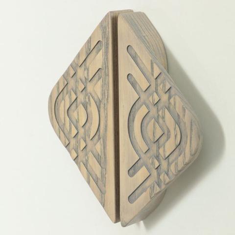 A wooden knob which consists of two symmetrical pieces that form a diamond shape when placed together. Each piece has intricate carvings with intersecting lines and shapes, creating a visually appealing design. The wood has a light, natural finish with a slightly weathered look.