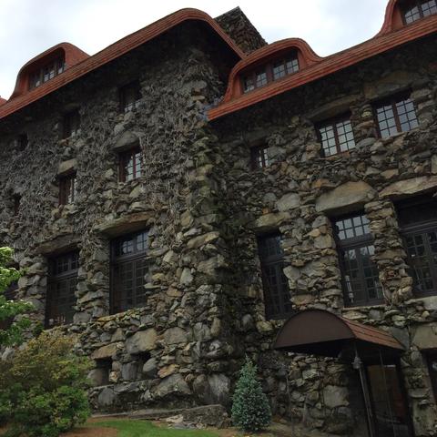Stone building with rustic architecture, featuring multiple windows and a distinctive red roof. Lush greenery surrounds the structure and winter-dead vines can be seen crawling up the stone.