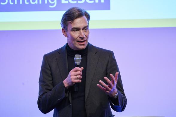 Ein Mann steht mit einem Mikrophon auf einer Bühne und spricht.