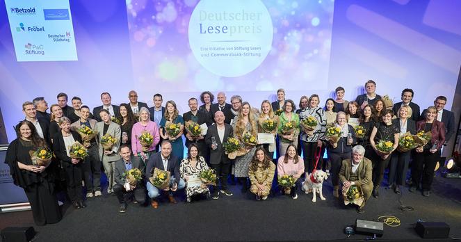 Auf einer Bühne stehen viele Menschen, die einen Blumenstrauß in der Hand halten. Im Hintergrund steht auf einer Leinwand: Deutscher Lesepreis