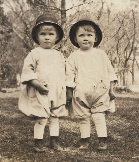 Close-up on the two boys... 1920 photo of two toddlers wearing rounded caps and big, puffy playclothes. Their tall white socks are easily visible above their old-fashioned playshoes. In the background, there are lots of trees, with few leaves on them. It is likely early Spring or early Autumn in northern Illinois, where the photo was taken. 

In the days before Pampers, Velcro, stain removers, colorfast clothing, and disposable diapers... Mom needed to be able to change their diapers easily and bleach dirty clothes. The solution was loose-fitting "Rompers" for playtime clothing.
