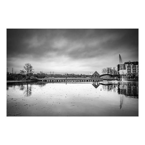 Paysage en noir et blanc capturant un plan d’eau calme sous un ciel nuageux. Un pont en bois traverse la scène, menant vers une petite maison au toit pentu. À droite, une fontaine projette un jet d’eau vertical, tandis que des immeubles se reflètent à la surface du lac, ajoutant une touche urbaine à cette atmosphère paisible.
