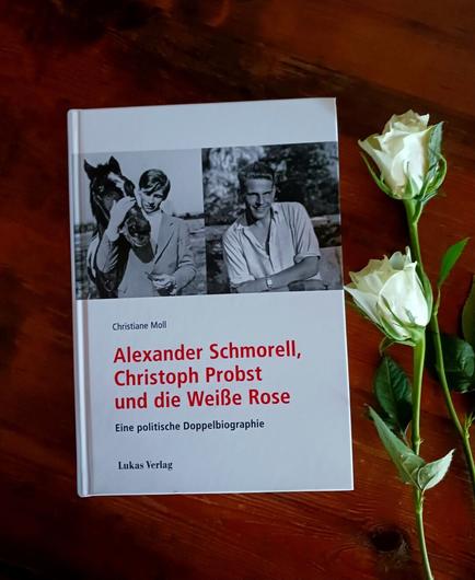 Foto des Buches, auf dessen weißem Cover zwei Schwarz-Weiß-Fotos von Alexander Schmorell mit einem dunklen Pferd (links) und Christoph Probst in offenem weißem Kurzarmhemd (rechts) zu sehen sind. Das Buch liegt auf dunklem Holz, rechts daneben liegen zwei weiße Rosen.