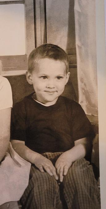 A photo of the same boy, 1964/5. Black shirt, checked pants. White curtain to his left, back of couch behind him.
