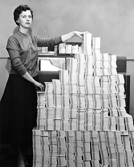 A 1960s black-and-white photograph of a woman with short hair dressed in contemporary fashion (a sweater with partially rolled up sleeves, long dark skirt) standing next to a pile of 62,500 punched cards neatly stacked in rows, one of her hands resting on the row closest to her, the other on the tallest, middle row.