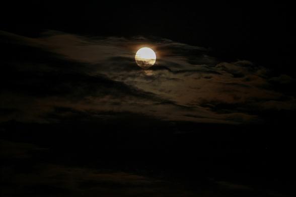 The image shows a full moon partly obscured by dark clouds in the night sky. The light from the moon illuminates the surrounding clouds, creating a dramatic and atmospheric effect.