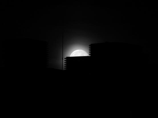 The image shows a black and white scene of a moon rising or setting behind silhouetted buildings. The moon is partially visible, appearing bright against the dark sky, with the buildings creating distinct outlines in the foreground.