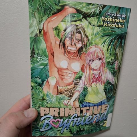 Photograph of a white person's hand holding Volume 2 of Primitive Boyfriend against a light colored wall. The cover shows a teenage girl in a Japanese school uniform in a very lush jungle next to a nearly naked ancient man who is pulling back some branches.