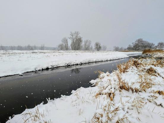 Snow-covered landscape with a dark river flowing through it, trees in the background, during a snowfall.