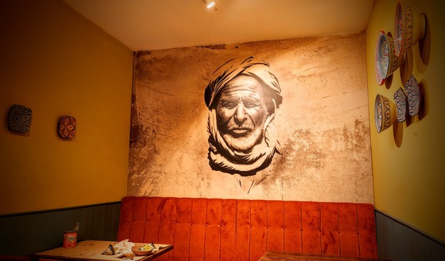 In einem kleinen Restaurant sieht man eine Sitzbank im American-Diner-Stil und davor Tische. An den Wänden hängen Töpferwaren mit andalusischen Mustern und auf einer Wand ist ein Mann als Wandbild aufgemalt, der zum Stamm der Tuarek gehören könnte.