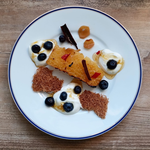 Das fertige Dessert auf dem Teller
Ein großes Stück gegrillte Ananas, 2 Stückchen Kadaifi-Knusper, 3 Kleckse Joghurt mit Zitronensirup einigen Stückchen Chili, Ingwer Zimtstange und Nelken und einige Heidelbeeren.