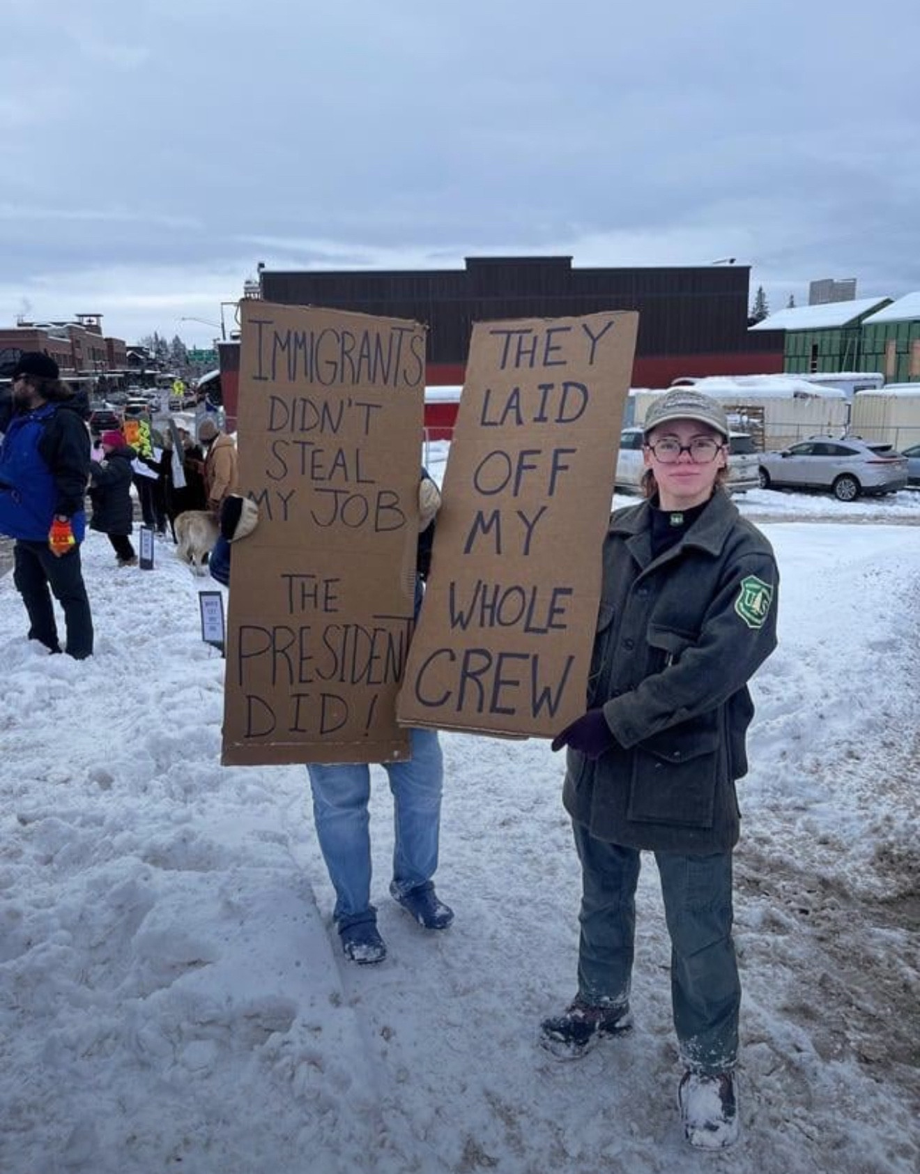 Image of people protesting in the snow, someone wearing USFS uniform Cardboard sign IMMIGRANTS DIDN'T STEAL MY JOB THE PRESIDENT DID! THEY LAID OFF MY WHOLE CREW