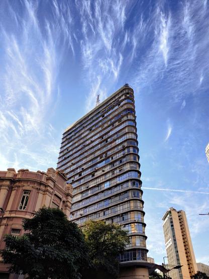 A tall, curved building, the Edifício Joaquim de Paula, dominates the center of the image, its facade reflecting the blue sky. The building is viewed from a low angle, emphasizing its height. The sky above is a vibrant blue with dramatic, wispy white clouds. To the left of the Edifício Joaquim de Paula, a smaller, more ornate building, part of the UAI Praça Sete complex, is visible. Other, more modern buildings are visible in the background. There are some trees in the lower part of the image.