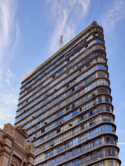A tall, curved building, the Edifício Joaquim de Paula, fills most of the frame, its facade reflecting the blue sky. The building is viewed from a low angle, emphasizing its height. This image is zoomed in, showing more detail of the building's windows and balconies. The sky above is blue with some wispy white clouds. A small portion of a more ornate, classical-style building, part of the UAI Praça Sete complex, is visible on the lower left.