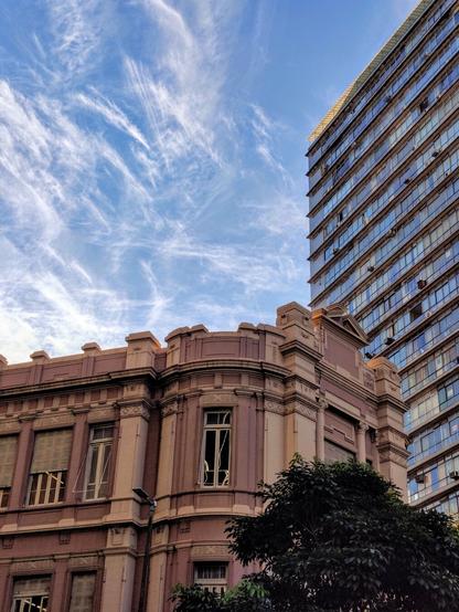 A low-angle shot showing two contrasting buildings. On the left, a shorter, ornate building with a pink and cream facade, part of the UAI Praça Sete complex. On the right, a portion of the tall, curved, modern Edifício Joaquim de Paula, with its reflective windows. The sky is blue with wispy white clouds. Green tree foliage is visible in the lower right corner of the frame.