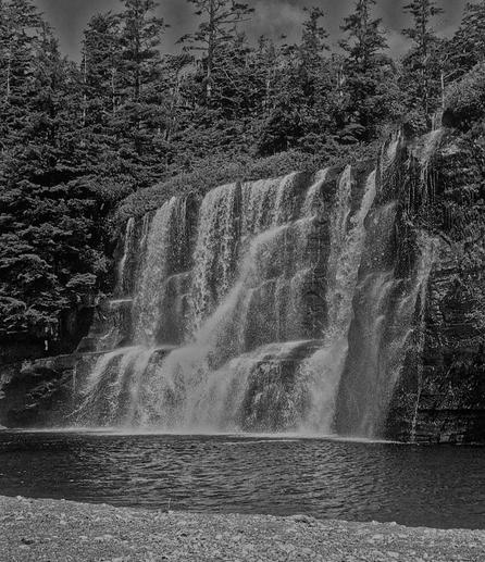 A black & white photo of a waterfall cascading over rocky cliff ledges into a calm pool below, surrounded by dense trees above. The water flows in multiple streams, creating a veil-like appearance. Beach rocks & sand gravel line the foreground.