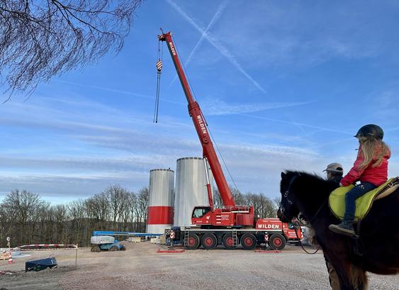 Baustelle, eine WKA wird errichtet. ein leuchtend roter Kran vor blauem Himmel dominiert das Bild, man kann Teile der Außenhülle des Mastes erkennen - und im Vordergrund ein braunes Pony mit Kind auf dem Rücken - beide betrachten die Szenerie.