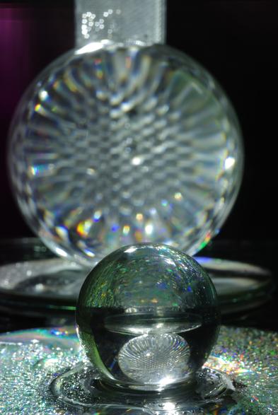 A reflector was placed behind a large glass ball on a CD, which reflected it in a point reflection. This ball is reflected in a point reflection by a smaller one on another wet CD. The CDs cause the LED light to be refracted, creating rainbow colours.