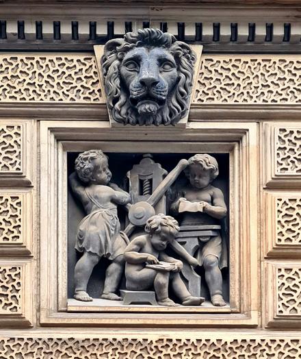 A sculpture of three putti printing bank notes on a printing press with a lion's head on the keystone above them.