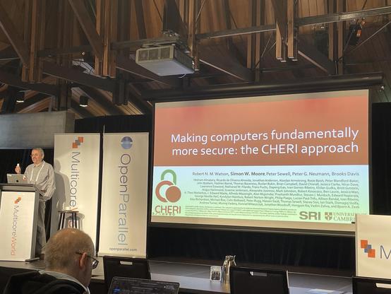 A speaker stands at a podium in front of a presentation slide titled "Making computers fundamentally more secure: the CHERI approach." The setting is a conference with banners for "Multicore World" and "OpenParallel."