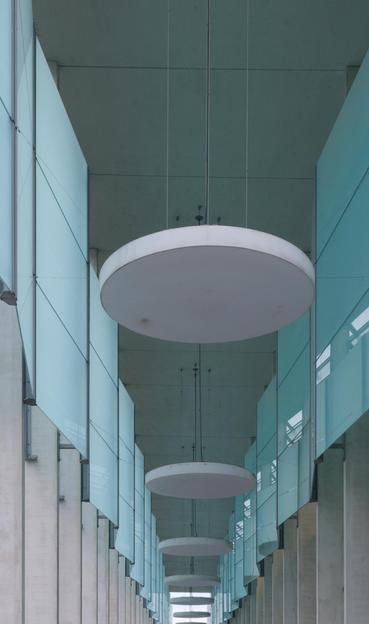 The image shows a perspective view of a modern architectural hallway. It features large, circular ceiling lights hanging from the ceiling, vertical concrete columns, and glass panels or walls on both sides. The image has a symmetrical composition, emphasizing the repetitive pattern of the architectural elements.