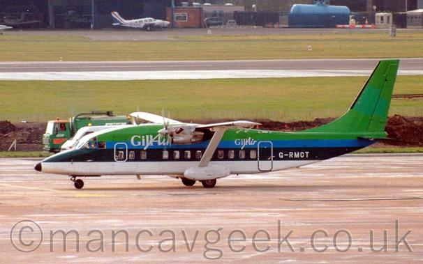Side view of a green, blue, and white, high-winged, twin propellor-engined airliner taxiing from right to left.
The plane is a vivid green on the top of the fuselage, a blue strip below that, a thick dark green stripe along the passenger cabin windows, and a white lower fuselage, with large, white "GillAir" titles on the upper forward fuselage, with a smaller version of this logo on the rear fuselage, just forward of the main door.
Grey concrete apron fills the foreground, with grass in the background leading up to a black runway, and more grass beyond that to a building with light aircraft parked outside in the distance.