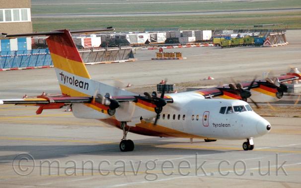 Side view of a white, hi-winged, 4 propellor-engined airliner taxiing from left to right.
The plane has thick dark umber, red, and yellow diagonal stripes running along the top of the tail and the front of the top of the engine pods, with the same stripes but in a reversed order running down the base of the tail and rear fuselage, curving under the body in front of the wing.
There are large black "Tyrolean" titles on the white part of the tail, repeated in a smaller form under the cockpit windows.
Grey taxiway fills the bottom of the frame, with more taxiways bordered by baggage carts and large luggage containers in the background, with acres of green grass leading off to the distance at the top of the frame.