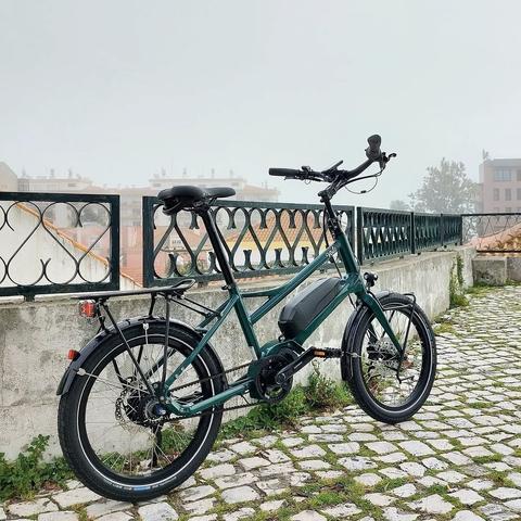 Bicicleta verde eléctrica, de rodas pequenas junto a um muro. Ao fundo telhados de casas e nevoeiro